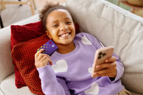 Girl lying down on a sofa holding a NatWest Adapt debit card and a phone.