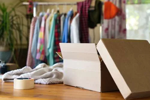 Open cardboard box on a wooden table with folded clothes nearby and a clothing rack in the background