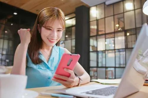A woman smiling while looking at her phone next to a laptop