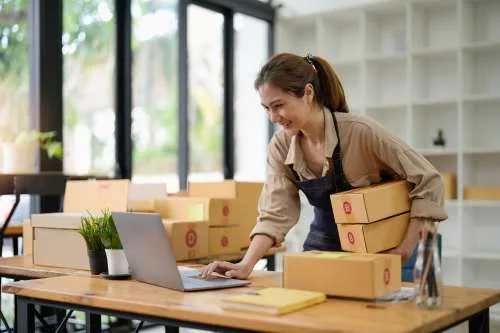 A small business owner packing boxes while working on a laptop