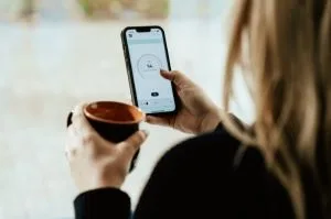A person holding a coffee mug while checking a smart home application on their smartphone.
