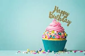 A colorful cupcake with pink frosting, rainbow sprinkles, and a gold glitter "Happy Birthday" topper against a light blue background.