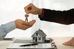 A real estate agent or seller handing over house keys to a buyer across a desk with a signed contract and a small model house.