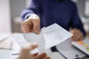 A person handing over a physical check to another person in a professional office setting.