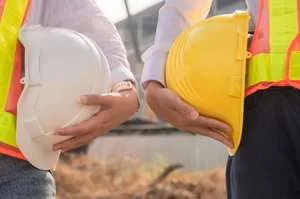 Two workers in high-visibility vests standing side-by-side, each holding a hard hat (one white, one yellow) under their arm.