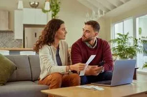 A young man and woman sit together on a grey sofa in a bright living room, looking at each other while discussing a paper bill.