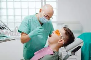 A dentist in green scrubs and a face mask performing a dental examination on a patient.