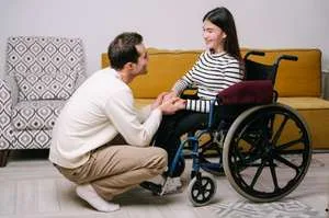 A young man kneels compassionately in front of a young woman in a wheelchair, holding her hands while they smile at each other in a bright living room.