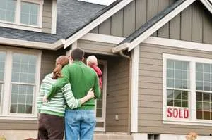 A back-view of a young family&mdash;a mother, father, and small child&mdash;standing together and looking at their newly purchased house.