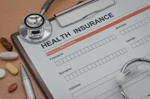 A health insurance application form on a clipboard, surrounded by a stethoscope, a pen, and various pills.