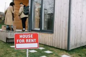 A suburban two-story house with a red "FOR RENT" sign displayed in the front window.