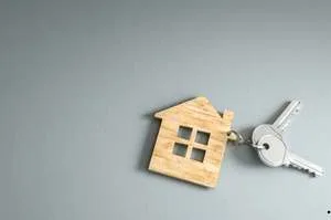 A pair of silver metal keys attached to a light wooden keychain shaped like a small house, lying on a solid grey background.