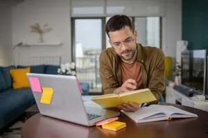A man with glasses sitting at a table in a home office, looking at a notebook while working on a laptop covered in colorful sticky notes.