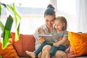 A smiling mother and her young daughter sit together on a bright, sunny couch looking at a digital tablet.
