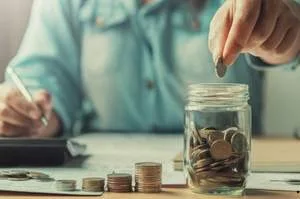 A person putting a coin into a glass jar filled with money, with stacks of coins and a calculator on a desk in the background.