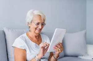 A smiling senior woman with glasses sitting on a grey sofa, comfortably using a white digital tablet.