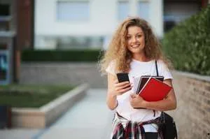 A smiling young woman with curly hair standing outside, holding textbooks and a smartphone, representing a student on campus.