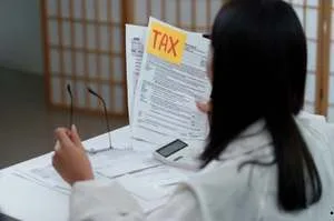 A person&rsquo;s hands organizing a stack of "Personal Income Tax" documents on a wooden desk next to a laptop and a calculator.