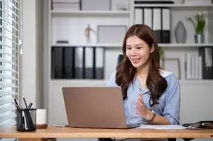 A smiling woman sitting at a desk and looking at a laptop during a video call in a bright office.