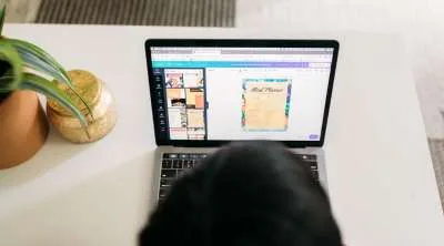 An over-the-shoulder view of a person using a laptop to design a "Meal Planner" document using an online graphic design interface.