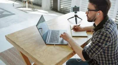 A man with glasses and earphones working on a laptop at a wooden desk, taking notes in a sketchbook with a smartphone on a tripod nearby.