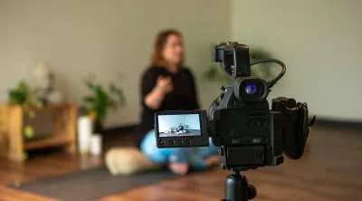 A professional video camera on a tripod in the foreground, filming a woman sitting cross-legged on a yoga mat in a bright room.