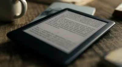 A close-up of an e-reader displaying a page of text, resting on a wooden surface next to a white ceramic mug.