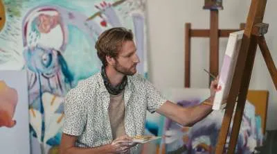 An artist with a beard and patterned shirt working on a canvas in a studio filled with large, colorful abstract paintings.