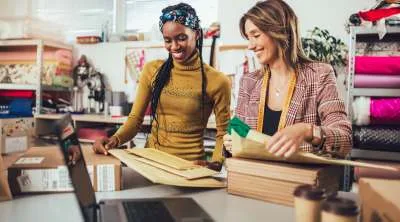 A woman in a warehouse setting carefully packing a cardboard box with tissue paper, preparing an order for shipping.