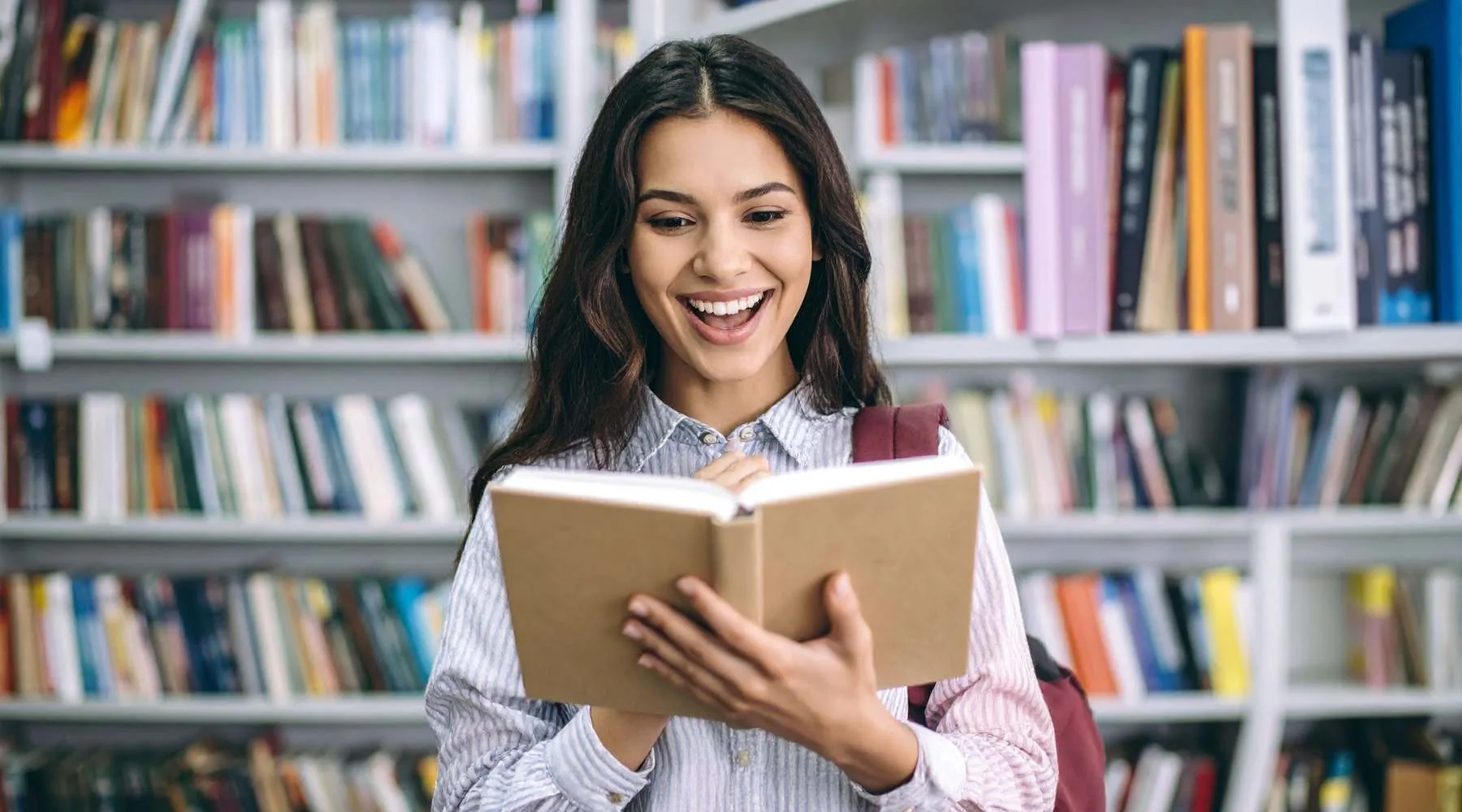 A smiling young woman holding an open book and looking delighted, with rows of bookshelves in a library blurred in the background.