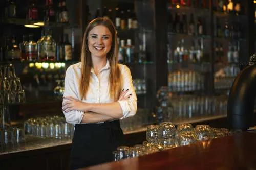 A young, smiling female bartender stands with her arms crossed behind a wooden bar.