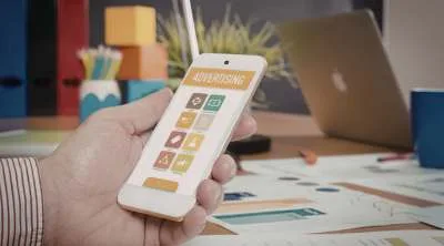 A hand holding a smartphone against a background of colorful, glowing digital icons like "Like," "Share," and "Chat," representing social media and digital marketing.