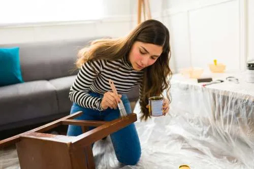 A young woman in a striped shirt and blue jeans kneels on a plastic-covered floor, carefully applying wood stain to a small wooden table with a paintbrush.