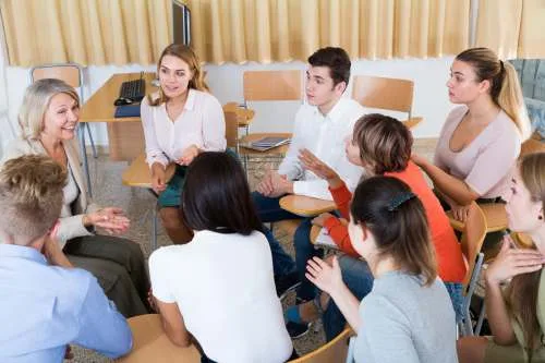 A group of diverse people sit in a circle of wooden desks in a bright room, engaged in an active discussion.