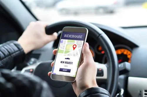 A close-up shot of a driver's hand holding a smartphone inside a car. The screen displays a "Rideshare" app