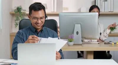 A person in front of a computer, using headphones and checking some papers.